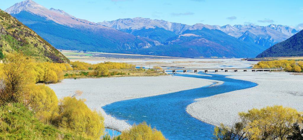 The braided Waimakariri River winding through a wide shingle riverbed in the Arthur's Pass valley in autumn, with golden willow trees lining the banks and the Southern Alps rising steeply on both sides.
