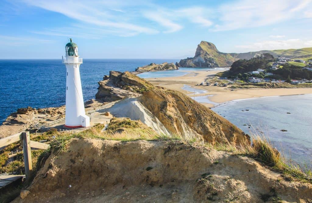 The white Castlepoint Lighthouse standing on a rugged limestone reef against the ocean under a clear blue sky.