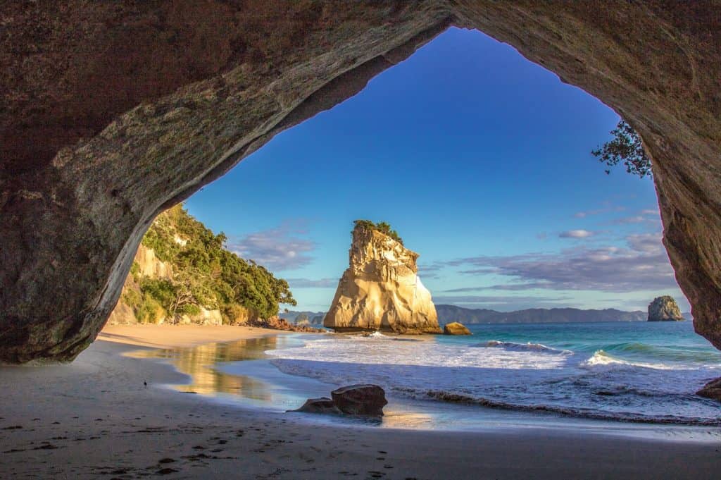 Looking through the natural limestone arch of Cathedral Cove toward a golden sand beach and dramatic rock stack rising from turquoise water, Coromandel Peninsula, New Zealand.
