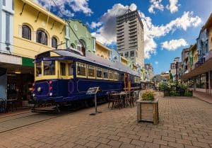 The restored blue and gold Christchurch heritage tram passing through the colourful pedestrian precinct of New Regent Street in the Christchurch CBD, with pastel-painted Spanish Mission style shopfronts and outdoor café tables lining the brick-paved street.