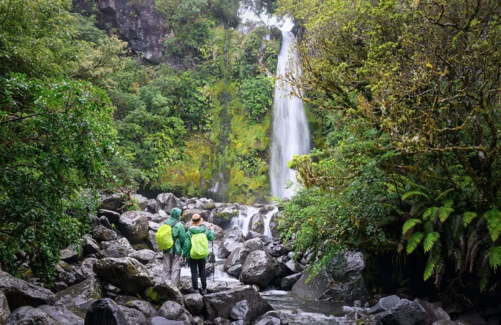 Two hikers in rain jackets standing on boulders at the base of Dawson Falls on the southern slopes of Mount Taranaki, the waterfall dropping through dense native rainforest into a rocky pool.
