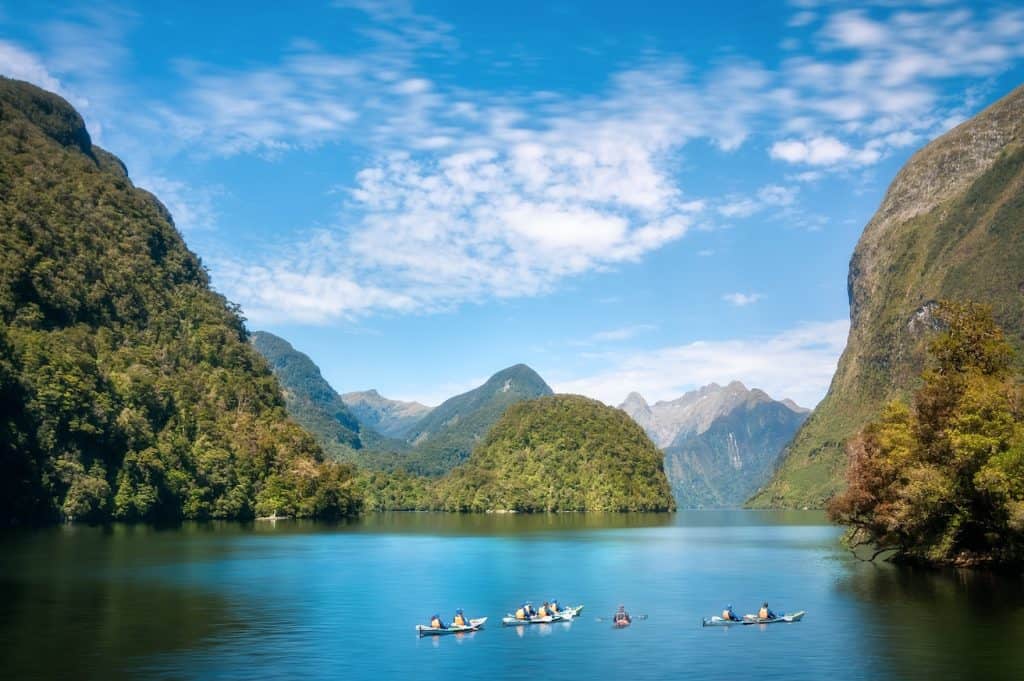 A group of sea kayakers paddling in formation across the glassy green waters of Doubtful Sound, surrounded by steep bush-covered fiord walls and layered mountain ranges receding into the distance under a blue sky with scattered cloud.
