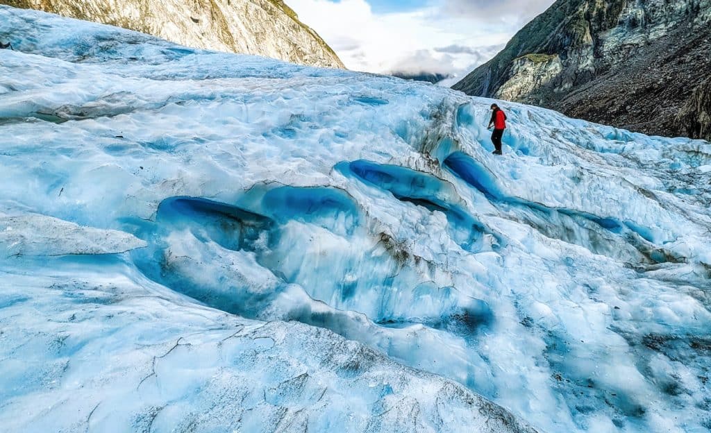 A lone hiker walking across the textured, bright blue ice of a massive glacier valley, surrounded by steep, rugged mountain rock walls under a slightly overcast sky.