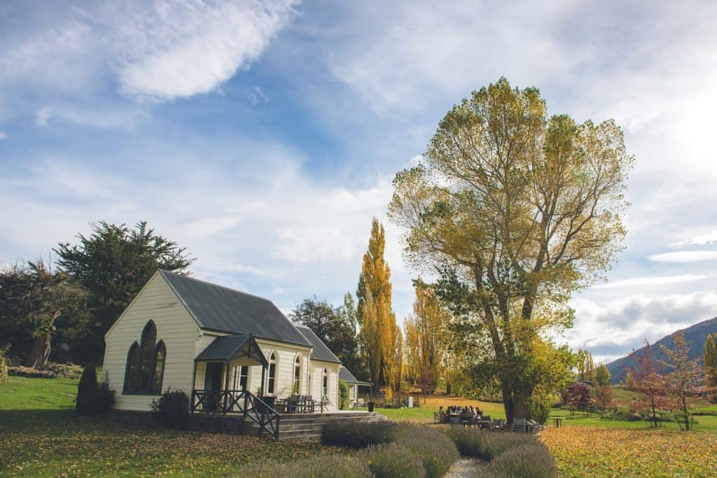 A historic white wooden chapel building used as a cellar door at a Gibbston Valley winery in autumn, surrounded by golden poplar trees, lavender borders, and guests dining on the lawn with the Central Otago ranges rising in the background.
