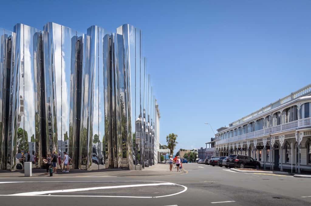 The mirrored stainless steel facade of the Len Lye Centre in New Plymouth reflecting the surrounding streetscape and sky, with pedestrians stopping to look at their distorted reflections, and a heritage building visible alongside.

