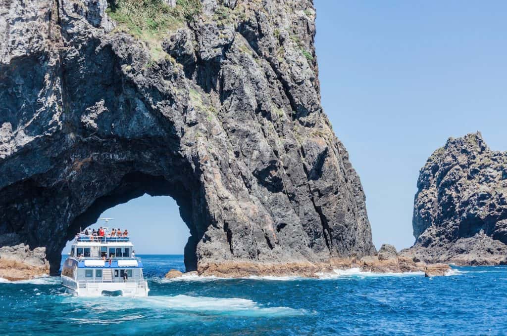 A sightseeing cruise boat navigating through the famous opening of the Hole in the Rock (Motukōkako) on a clear day in the Bay of Islands, New Zealand.
