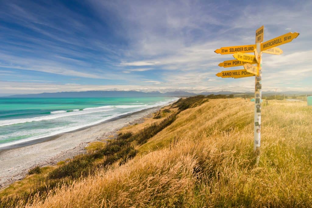 A yellow directional signpost pointing toward Solander Island, Invercargill, Codfish Island, and Sand Hill Point, standing above a sweeping Southland beach with turquoise surf, dry tussock grass, and the distant Fiordland ranges on the horizon.
