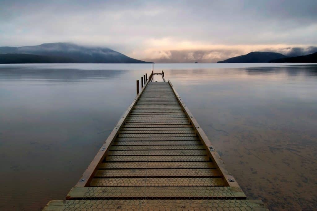 A wooden jetty extending into the vast, perfectly still surface of Lake Hauroko at dusk, New Zealand's deepest lake, with low cloud resting on the dark forested hills of the Fiordland wilderness rising from the far shore.