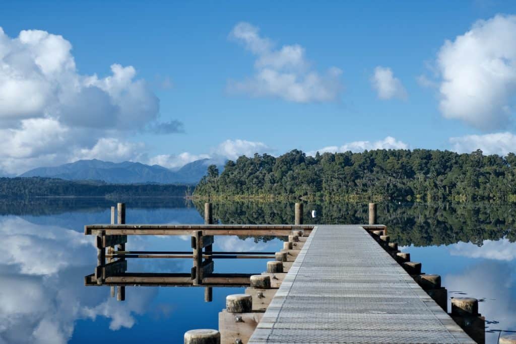 A wooden jetty extending into the mirror-still surface of Lake Mahinapua near Hokitika, reflecting a cloudscape and the dense native kahikatea forest lining the far shore, with the Southern Alps visible in the distance.
