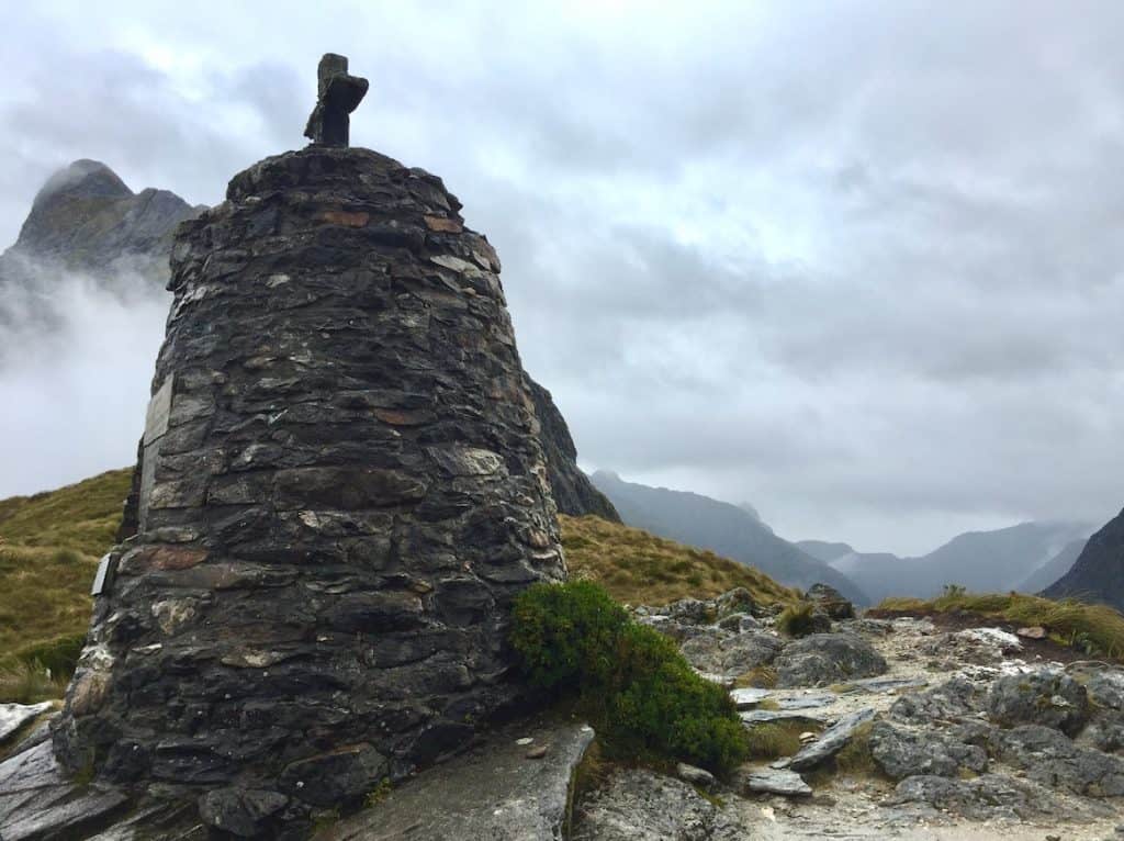The stone cairn memorial at MacKinnon Pass on the Milford Track at 1,154 metres, with low cloud wrapping the surrounding Fiordland peaks and tussock-covered alpine terrain stretching toward the valley below, Southland, New Zealand.
