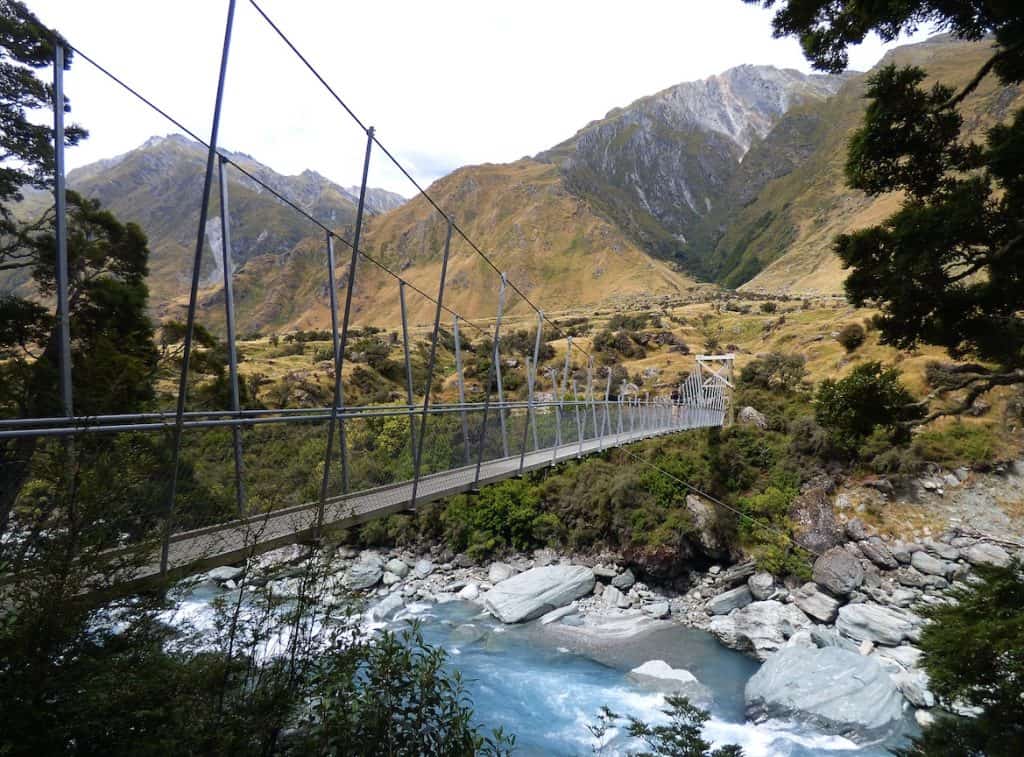 A swing bridge spanning a fast-flowing glacial stream on the Rob Roy Glacier Track in the Matukituki Valley, with the tawny tussock-covered slopes and rocky peaks of Mount Aspiring National Park rising steeply in all directions, Wanaka, New Zealand.