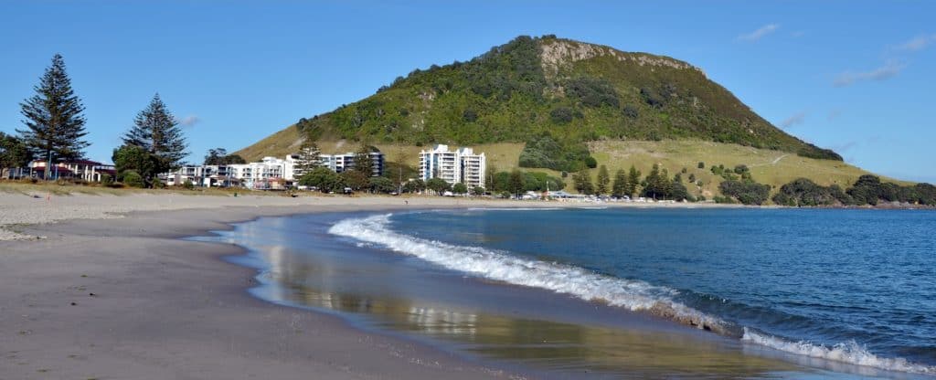 The sheltered beach on the Pilot Bay side of Mount Maunganui, with gentle waves lapping the shore and the green volcanic cone of Mauao rising steeply behind the beachfront apartments.