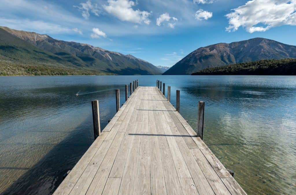 The iconic wooden jetty extending into the calm glacial waters of Lake Rotoiti, framed by the rugged, forest-clad mountains of Nelson Lakes National Park.