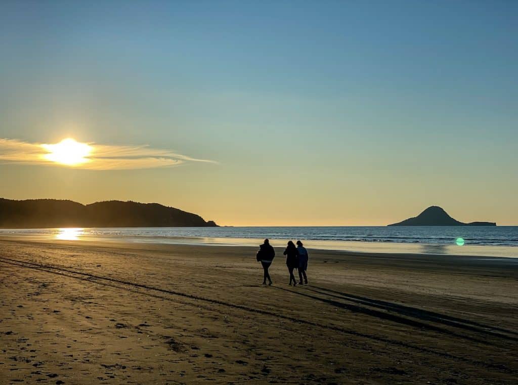Three silhouetted walkers on Ōhope Beach at sunset, with Whale Island visible on the horizon and a warm golden sky reflected on the wet sand, Whakatāne, Bay of Plenty.