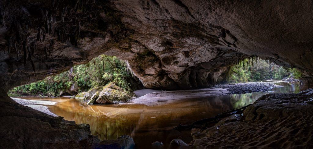 Looking out from inside the vast Oparara Arch in the Oparara Basin near Karamea, with the tannin-stained river flowing across a sandy floor beneath the massive limestone ceiling and ancient podocarp forest framing the exit.
