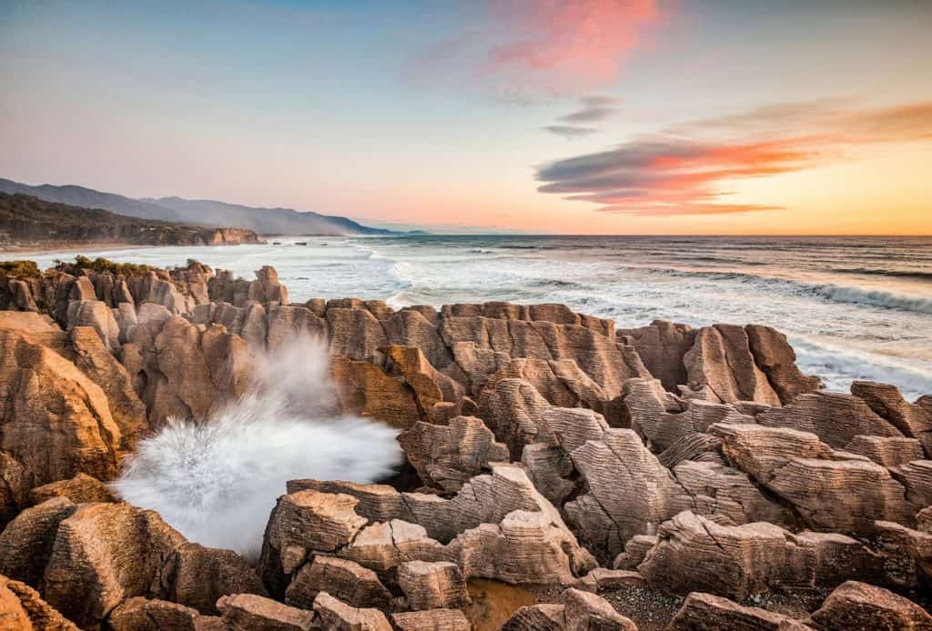 A blowhole erupting between the layered limestone Pancake Rocks at Punakaiki at sunset, with waves breaking against the West Coast shoreline and a vivid pink and orange sky above the Tasman Sea.