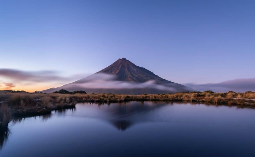 Mount Taranaki perfectly reflected in the still water of the Pouakai Tarns at dusk, with low cloud wrapping the volcanic cone's mid-slopes and golden tussock grass framing the foreground.
