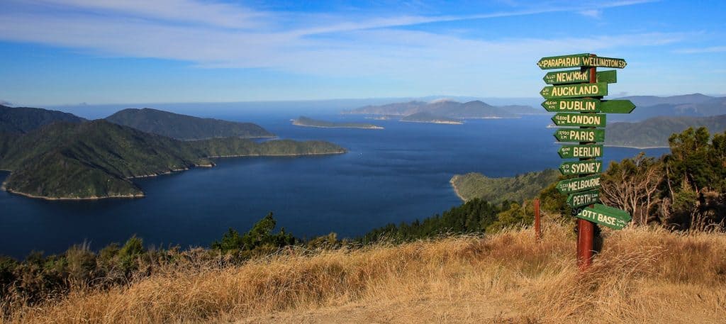 Panoramic view of Queen Charlotte Sound with a wooden distance signpost in the foreground, showing blue water, lush green hills, and the Marlborough Sounds landscape.