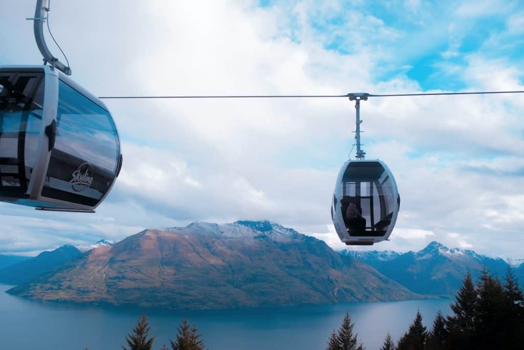 Two Skyline gondola cabins ascending above Queenstown on a cloudy day, with Lake Wakatipu and the snow-dusted peaks of the Cecil and Walter ranges stretching across the horizon below.
