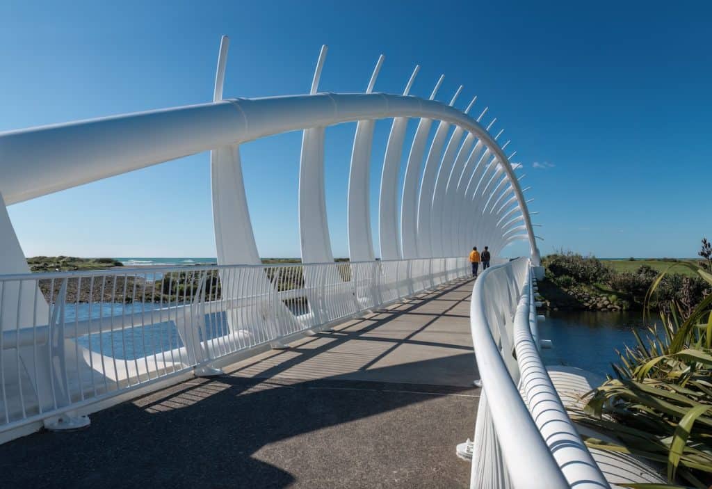 Two walkers crossing the striking white Te Rewa Rewa Bridge on the New Plymouth Coastal Walkway, its arching rib structure framing views of the Tasman Sea coastline and native coastal vegetation.
