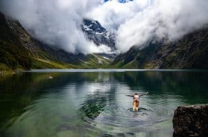 A swimmer wading into the crystal-clear glacial waters of Lake Marian in Fiordland, arms outstretched, with the steep bush-covered valley walls, snow-dusted peaks, and dramatic cloud formations of the Darran Mountains reflected in the still green water behind her.