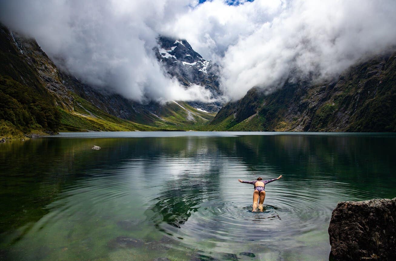 A swimmer wading into the crystal-clear glacial waters of Lake Marian in Fiordland, arms outstretched, with the steep bush-covered valley walls, snow-dusted peaks, and dramatic cloud formations of the Darran Mountains reflected in the still green water behind her.