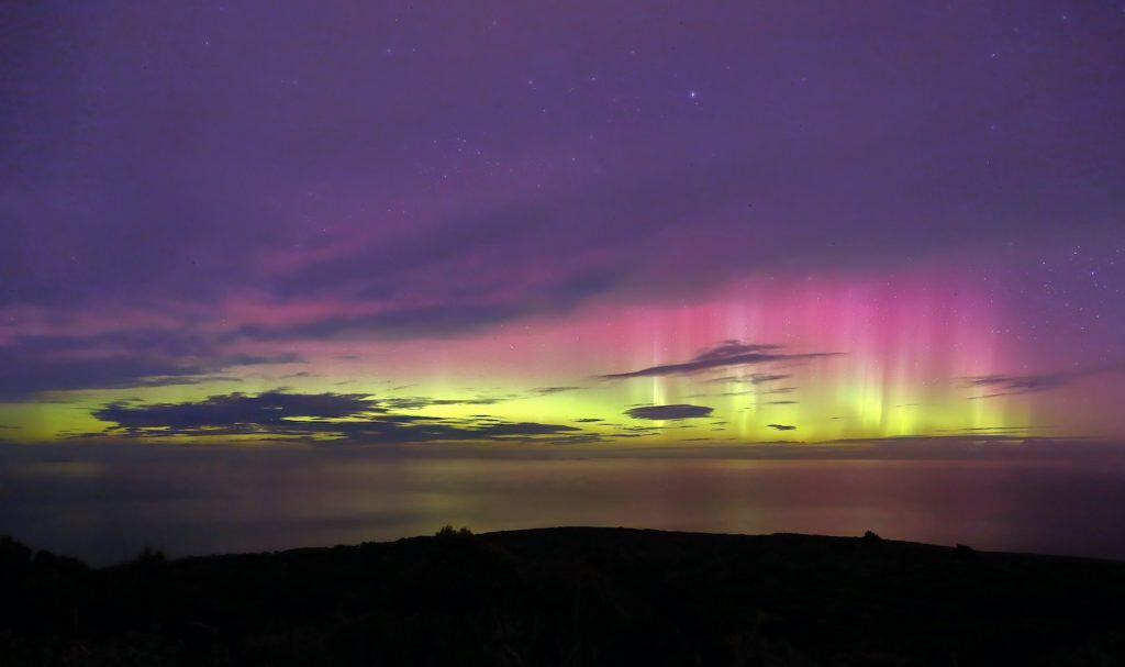 The aurora australis (Southern Lights) displaying vivid columns of pink, magenta, and yellow-green above the ocean horizon as seen from Stewart Island/Rakiura, with a dark silhouetted headland and scattered stars visible in the purple night sky.