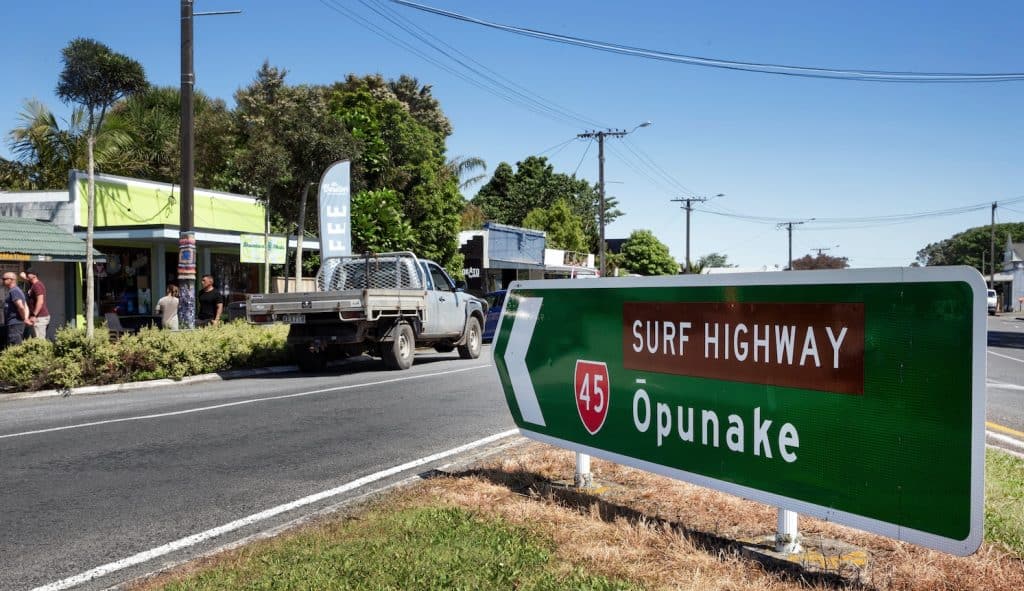 A green and brown road sign for Surf Highway 45 pointing toward Ōpunake on a sunny day, with a small coastal town and palm trees visible in the background, Taranaki.