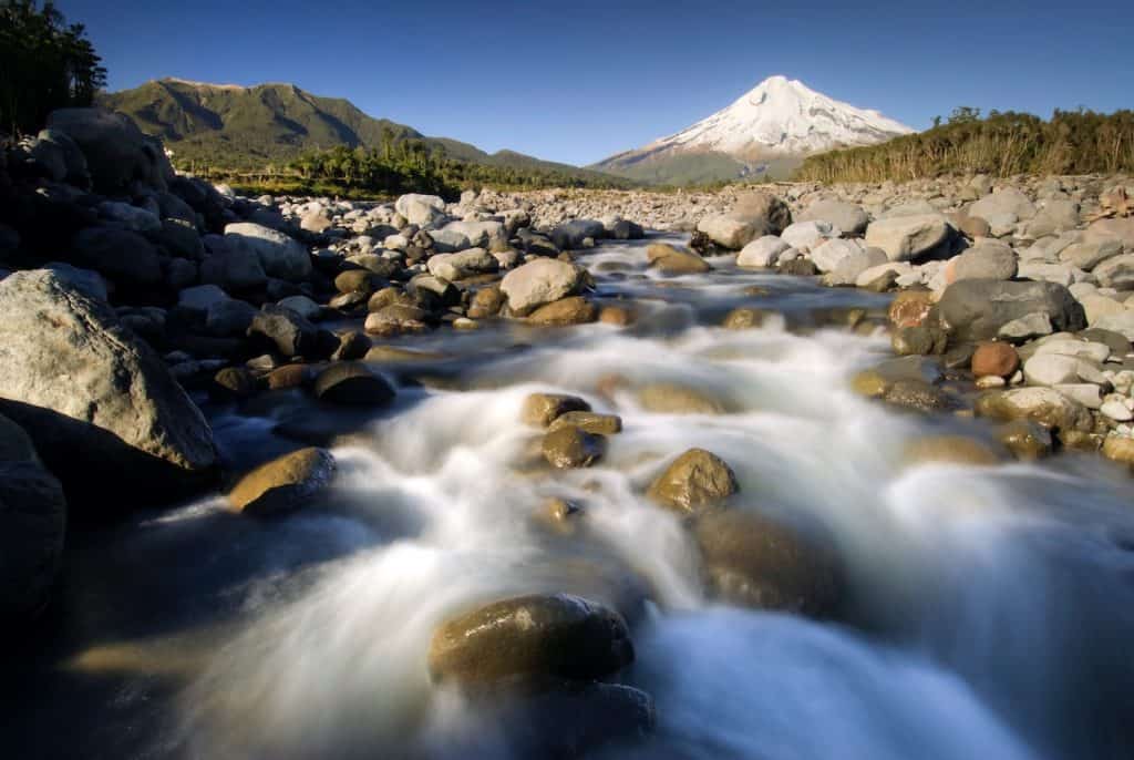 A fast-flowing boulder-strewn river on the lower slopes of Mount Taranaki, with the snow-capped volcanic summit rising sharply against a deep blue sky and native forest flanking both banks.
