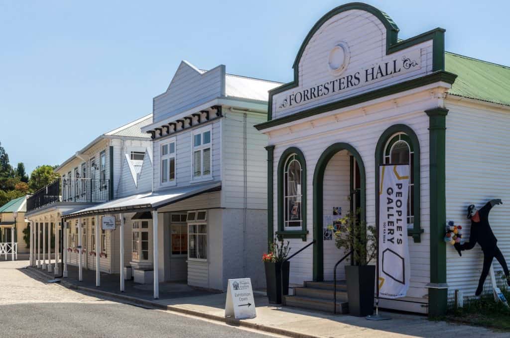 The heritage Forresters Hall building at the Tauranga Historic Village, housing the People's Gallery art exhibition, with its distinctive white timber facade and green trim under a clear blue sky.
