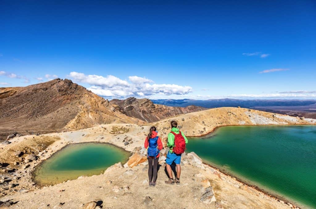 A couple with hiking backpacks walking along the volcanic trail of the Tongariro Alpine Crossing between the Emerald Lakes and Blue Lake, New Zealand.