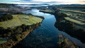 Aerial view of the Waipā River winding through frost-touched green farmland in the Waikato at dawn, with low morning mist hanging over the river flats and the ranges visible on the horizon.