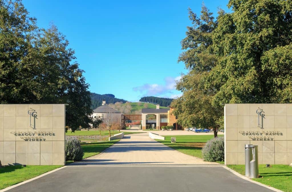 The grand entrance to the architecturally designed Craggy Range Winery buildings, framed by lush vineyards and the dramatic stone cliffs of Te Mata Peak rising in the background.