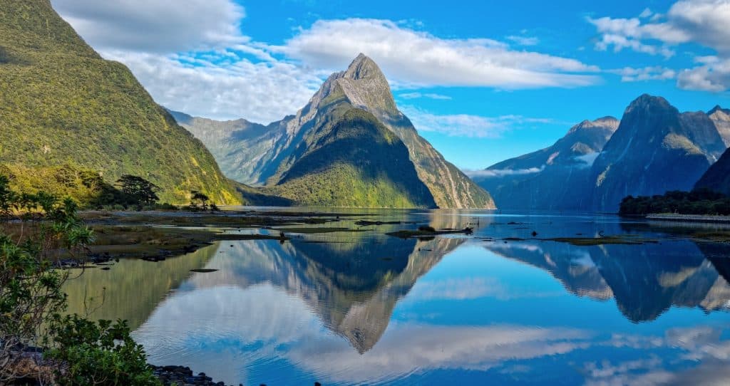 Mitre Peak rising 1,692 metres directly from the mirror-still surface of Milford Sound (Piopiotahi) on a clear blue morning, with the steep forested walls of the fiord and scattered cloud reflections perfectly mirrored in the water below.
