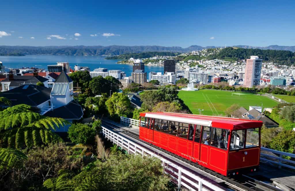 The historic red Wellington Cable Car ascending the hillside tracks with the city skyscrapers and harbour in the background.
